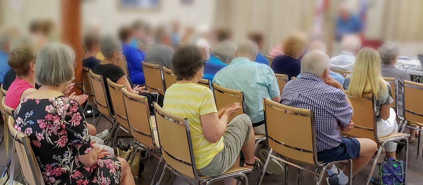 A community hall filled with residents seated for a public meeting, facing a speaker at the podium